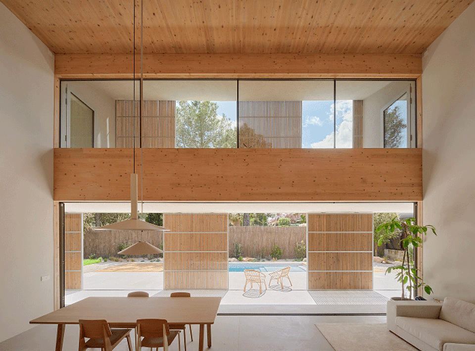 House in Girona with a double-height central space that fully opens onto the terrace and swimming pool. This house in Girona incorporates wooden filters and large openings to combine natural light, solar protection, and a strong connection with the outdoors