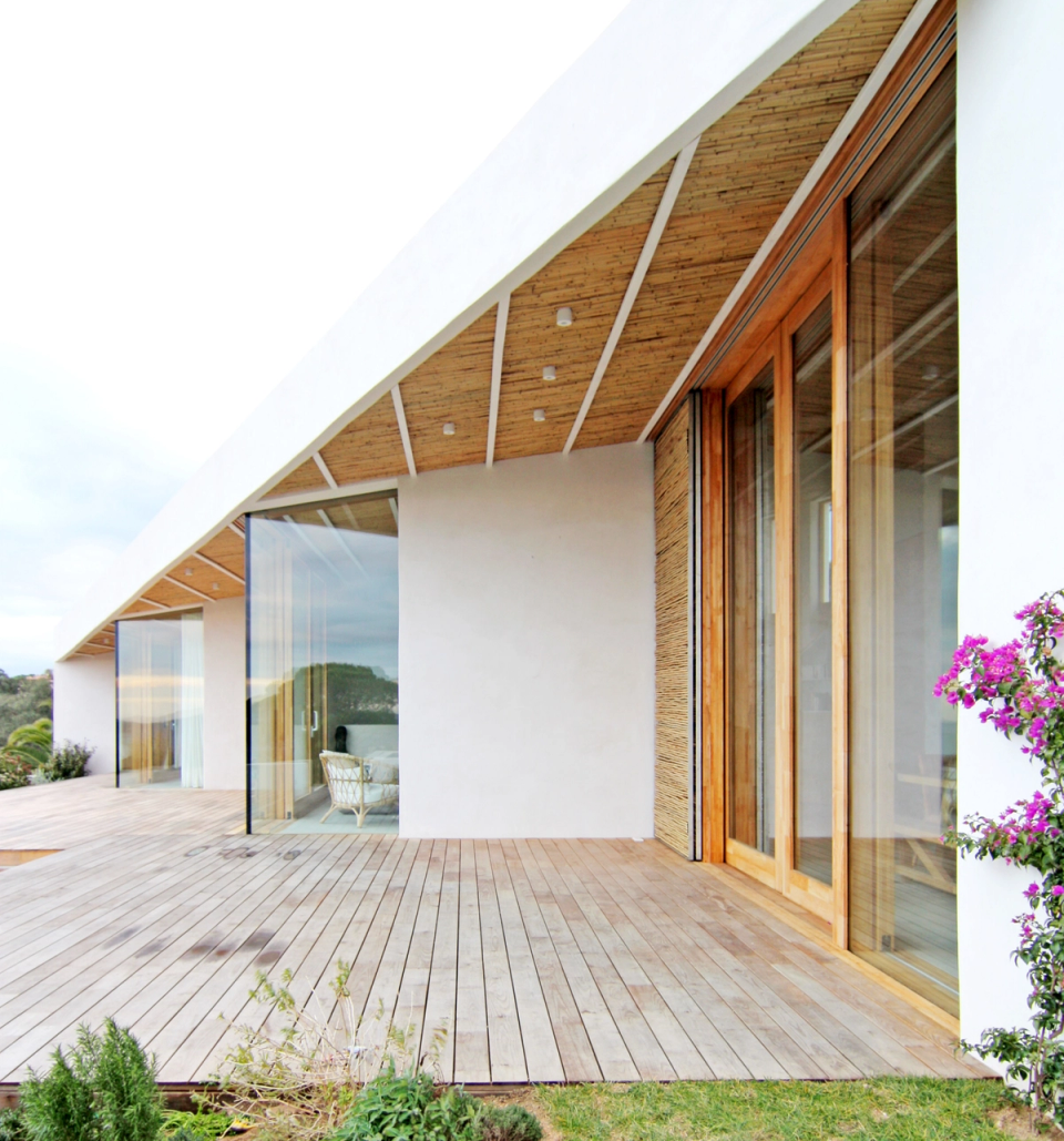 Detail of the outdoor terrace of a contemporary detached house with large glass enclosures, natural wood finishes and a pitched roof that adds dynamism and protection to the outdoor spaces. The combination of warm materials, clean lines and direct connection with the garden creates a bright and elegant atmosphere fully integrated with the Mediterranean surroundings.