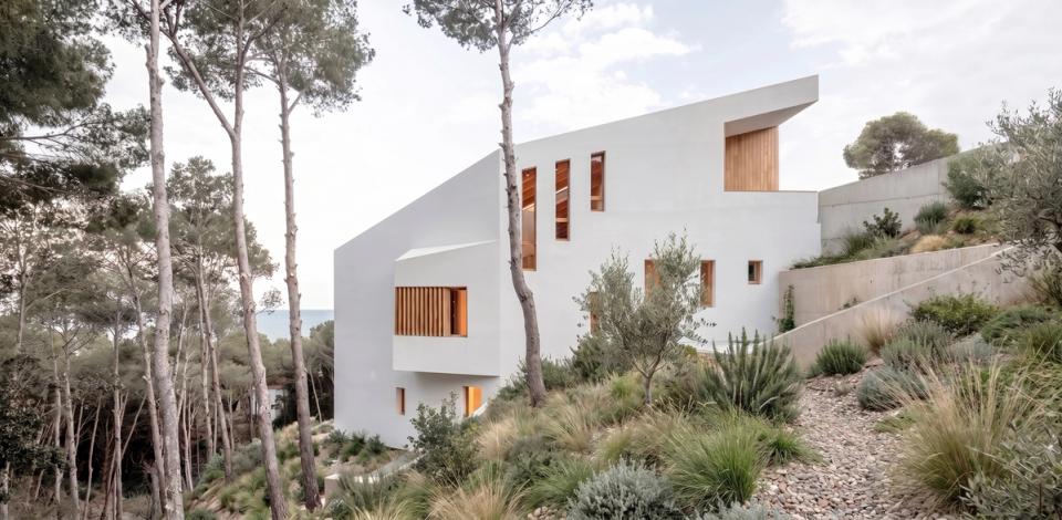 Contemporary detached single-family home delicately integrated into a Mediterranean forest setting. The project adapts to the sloping plot through sculpted volumetry and a layout that respects the topography and existing vegetation. White façades combined with vertical openings and wood details provide brightness, warmth, and an elegant timeless architectural identity. This detached house prioritizes the relationship with the landscape, privacy, and natural light, creating interior spaces visually connected to the outdoors and perfectly integrated into the surroundings.