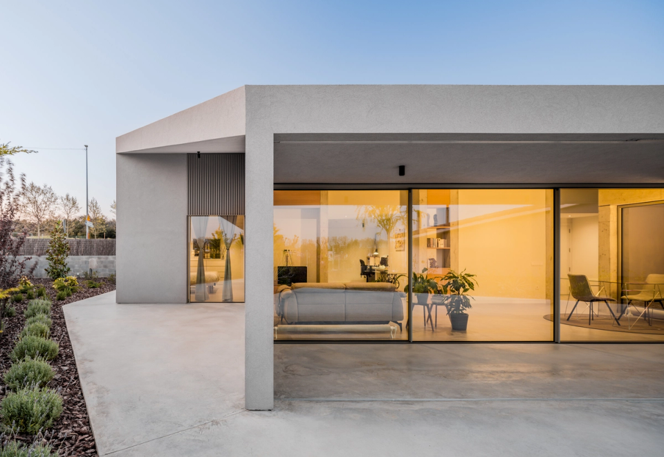 This single-family home stands out for its pure volumetry and the fluid connection between interior and exterior through large sliding windows. The minimalist composition, with continuous finishes and a neutral palette, reinforces the feeling of spaciousness and calm, while the warm interior lighting brings comfort and intimacy to the living spaces.
