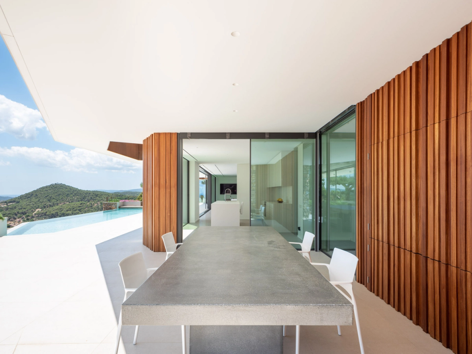 The detached single-family home is characterized by a contemporary architecture with very clean lines, featuring large white cantilevers, glass façades, and vertical wood cladding that brings warmth to the ensemble. The continuous terrace and infinity pool open completely to the landscape, creating a direct connection between interior and exterior through large sliding openings. The combination of natural materials, open spaces, and a neutral palette creates an elegant, bright atmosphere integrated with the Mediterranean surroundings.
