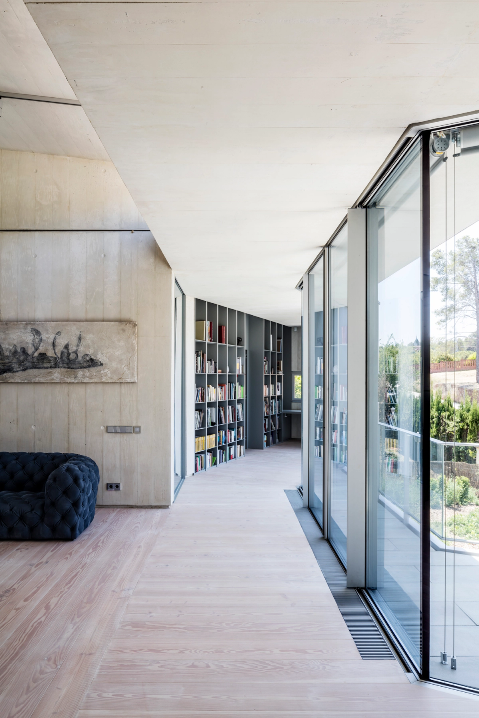 Interior corridor of a contemporary single-family home with an open and minimalist architecture that prioritizes natural light and visual connection between spaces. Large sliding glass openings integrate the exterior into the house, while continuous exposed concrete finishes, light wood, and integrated furniture provide coherence and elegance to the whole. The linear bookshelf at the end enhances the visual depth of the space and contributes to creating a serene, functional, and sophisticated atmosphere.