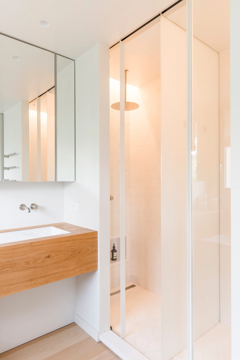 Interior renovation of a contemporary bathroom with a bright minimalist aesthetic dominated by white tones and natural wood finishes. The suspended wooden furniture adds warmth and texture to the space, while the integrated sink and wall-mounted faucet reinforce the sense of visual cleanliness and sophistication