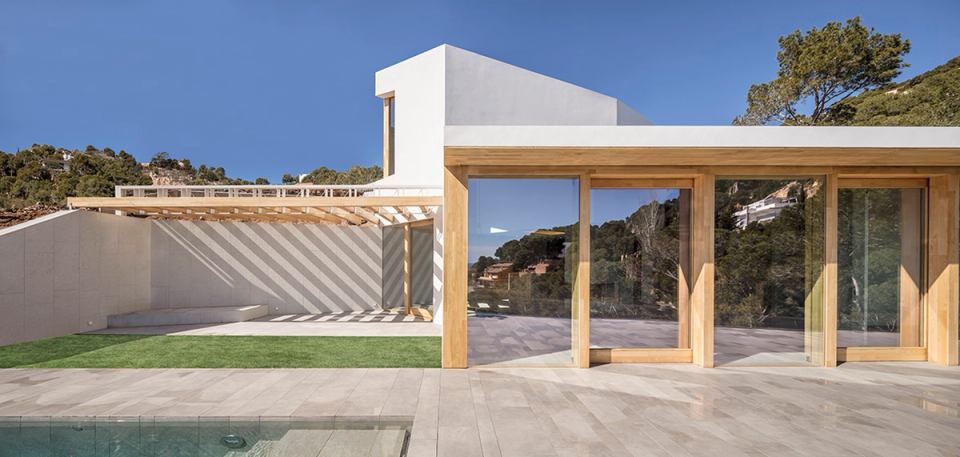 Detached single-family home with contemporary architecture featuring a strong presence of natural wood and large glazed surfaces that enhance the connection between interior and exterior spaces. The composition of pure volumes, the lightweight pergola, and the projected shadows add visual dynamism and reinforce the Mediterranean character of the house. The design prioritizes natural light, openness to outdoor spaces, and integration with the surroundings, creating a warm, functional, and elegant atmosphere designed to enjoy open-air living all year round.