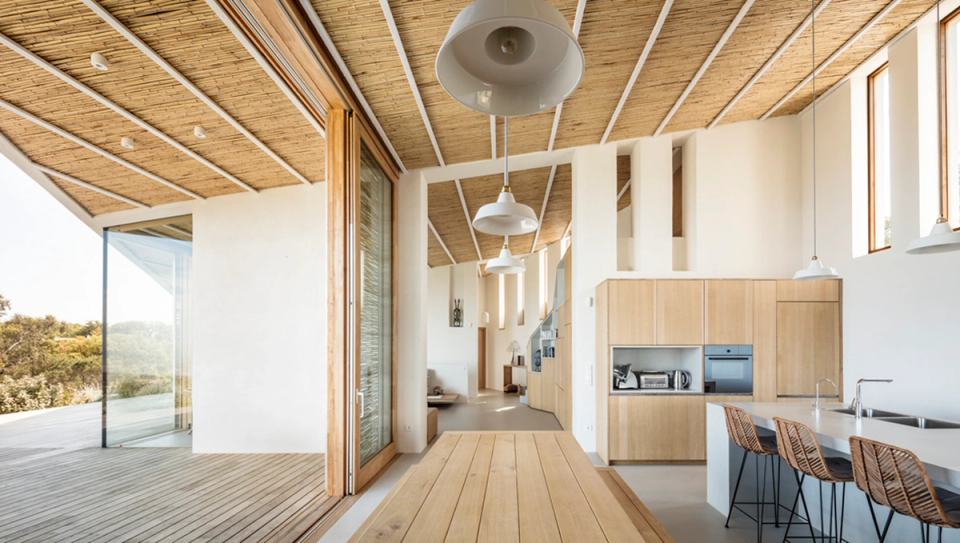 Dining room and open kitchen of a detached house with warm minimalist architecture defined by natural wood ceilings, large panoramic openings and simple-lined furniture. The interior space opens completely towards the terrace and outdoor landscape, enhancing natural light and the sense of continuity between the home, nature and the Mediterranean lifestyle.