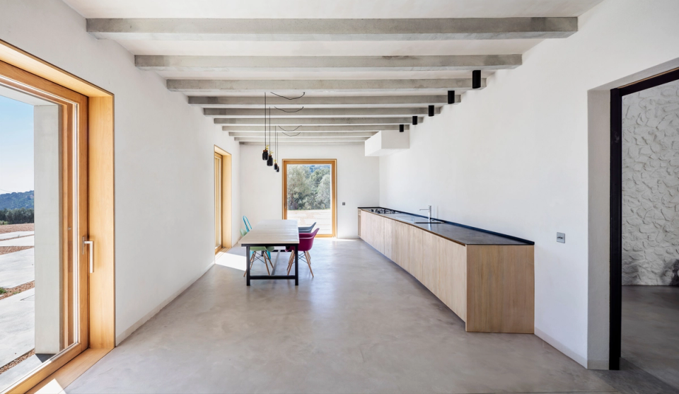 Bright interior of a comprehensive renovation featuring an open kitchen and open-plan spaces designed to enhance functionality and the connection with the outdoors. Exposed concrete beams, continuous flooring, and natural wood finishes add character and warmth to a contemporary and minimalist atmosphere. Large openings allow natural light to enter throughout the day, reinforcing the sense of spaciousness and comfort. This full renovation combines modern architecture, noble materials, and an open layout that encourages daily living and the relationship between spaces.