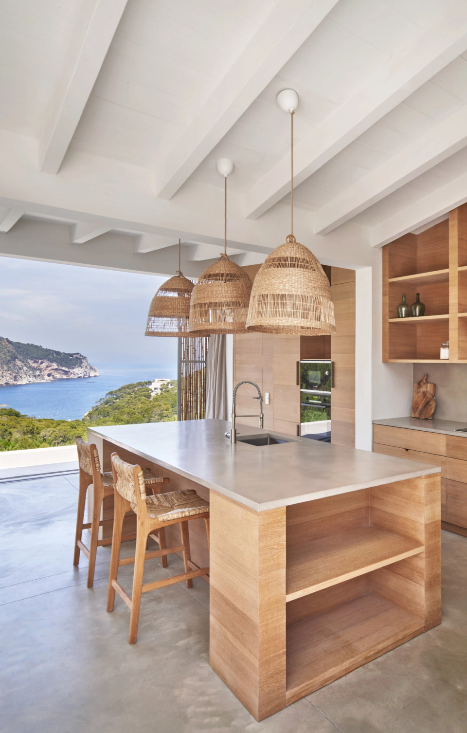 The kitchen of this single-family home integrates completely with the landscape thanks to large panoramic openings that connect the interior with sea views and the natural surroundings. The space combines a contemporary Mediterranean aesthetic with warm materials such as natural wood, plant fibers, and mineral finishes, creating a bright and welcoming atmosphere. The central island becomes the social core of the single-family home, providing functionality and visual continuity with the rest of the daytime living area. The combination of natural light, exposed beam ceilings, and neutral tones reinforces the sense of spaciousness and serenity characteristic of the architectural project.