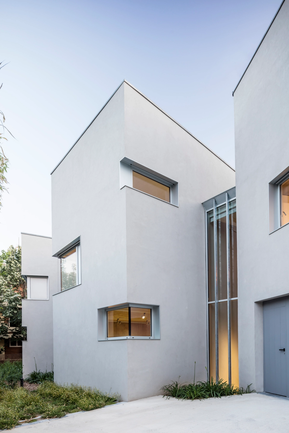 Minimalist single-family house with a composition of pure geometric volumes that define an elegant and functional contemporary architecture. The light-colored façades combine with large openings and corner windows that bring natural light into the interior spaces, while the glazed vertical access creates a sophisticated focal point and reinforces the visual connection between floors. The integration of vegetation and the simplicity of materials enhance the clean and timeless aesthetic of the residential project.