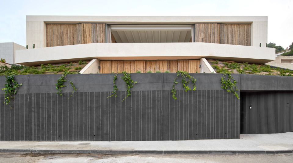 This single-family home presents a strong and balanced architectural composition, defined by horizontal lines that reinforce integration with the terrain and the Mediterranean landscape. The project combines a solid and dark base, intended for access and service spaces, with a lighter upper volume clad in natural materials that provide warmth and texture. The vegetation integrated into the embankments and walls helps soften the presence of the building and strengthen its relationship with the surroundings. This single-family home embraces contemporary, minimalist, and functional architecture, where privacy, natural light, and outdoor connection become key elements of the project.