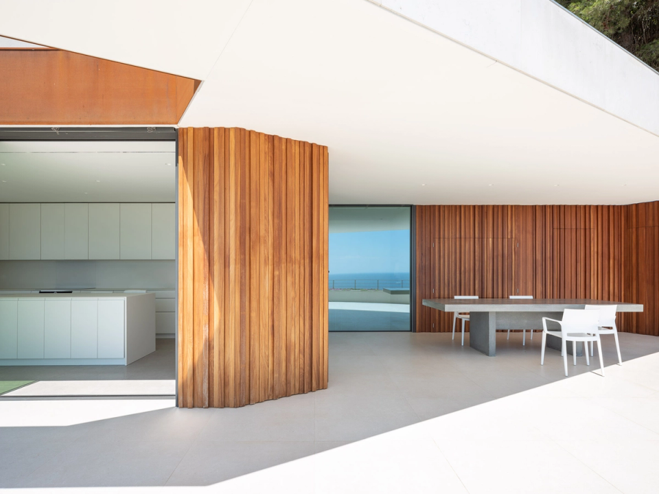 The detached single-family home combines contemporary clean-lined architecture with a strong connection to the landscape through large glass surfaces, white cantilevers, and vertical wood cladding that adds warmth. The continuous terrace and infinity pool visually merge with the horizon, while the interior spaces fully open to the outdoors through large-format sliding openings. The combination of light concrete, natural wood, and continuous flooring creates a minimalist, bright, and elegant atmosphere.