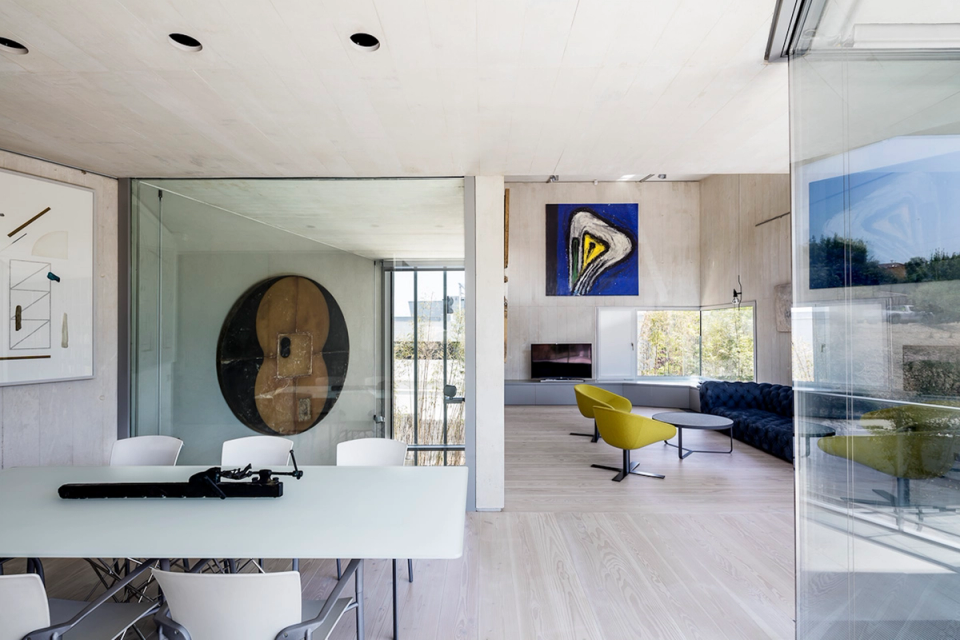 Interior of a contemporary single-family home with an open layout that connects different spaces in a fluid and functional way. Large windows and glass enclosures enhance the entry of natural light and create a constant relationship with the outdoors, providing spaciousness and transparency throughout the space. The use of neutral materials, continuous flooring, and industrial-style ceilings coexists with unique furniture and art pieces, shaping a modern, elegant atmosphere with a strong architectural identity.