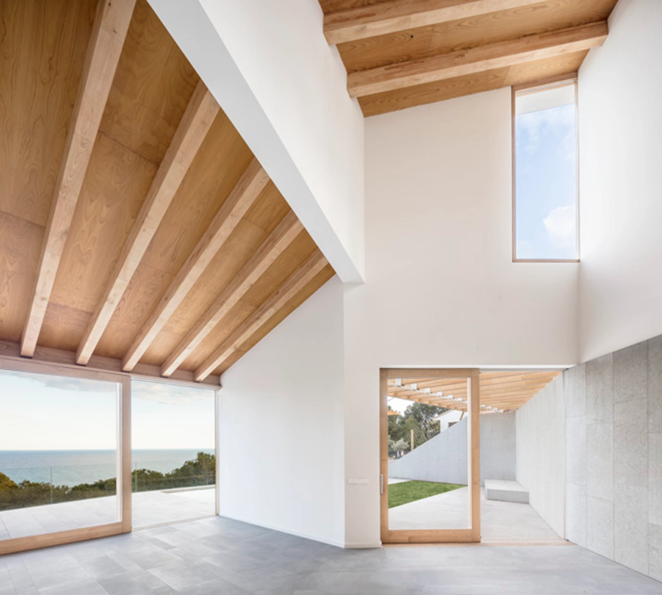 Interior space of a detached single-family home with minimalist lines and large open volumes, where sloped ceilings with exposed wooden structures provide warmth and visual rhythm. Large panoramic openings connect directly with the outdoors and offer views of the landscape and the sea, enhancing natural light and the fluid relationship between architecture and surroundings.