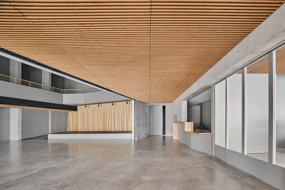 Interior of a cultural center with an open and flexible layout designed to adapt to stage activities, exhibitions, and social events. Continuous wooden slat ceilings provide acoustic comfort and a strong sense of warmth, while exposed concrete and neutral finishes reinforce the contemporary character of the space. The integrated stage, reception areas, and visual connection between levels turn this cultural center into a modern, functional, and versatile facility.