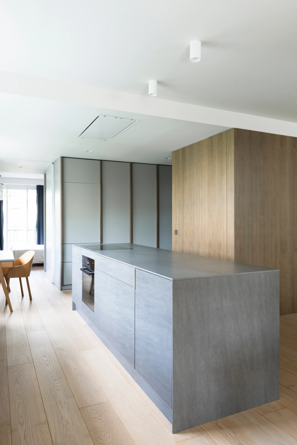 Interior renovation of a contemporary kitchen designed with an open and functional layout. The space combines a large central island with linear cabinetry in dark finishes and natural textures, creating an elegant and balanced atmosphere. The presence of wood adds warmth to the ensemble, while natural light filtered through large windows reinforces the sense of spaciousness and comfort. The project embraces clean lines, noble materials, and subtle spatial integration to achieve a minimalist and timeless aesthetic.