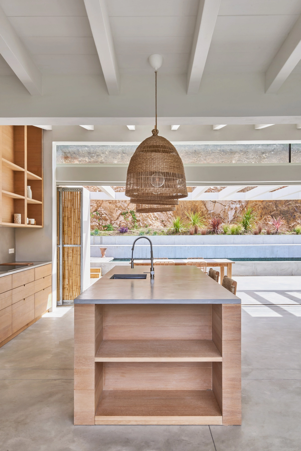 The kitchen area of this single-family home embraces open and fluid architecture, where interior and exterior spaces relate naturally. The large central island organizes the space and reinforces the kitchen’s functional and social character, while wood finishes and natural fiber lighting fixtures provide a warm and timeless Mediterranean aesthetic. Large openings allow constant cross ventilation and the entry of natural light, enhancing the feeling of spaciousness. This single-family home combines minimalism, noble materials, and landscape integration to create contemporary spaces with a strong connection to the outdoors.