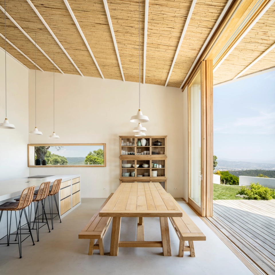 Living area of a detached house featuring an open kitchen, dining room and circulation spaces integrated into one spacious and bright environment. Natural wood ceilings, large sliding openings and minimalist furniture reinforce the sense of warmth and visual continuity, creating a seamless connection between the interior, the outdoor terrace and the surrounding natural landscape.