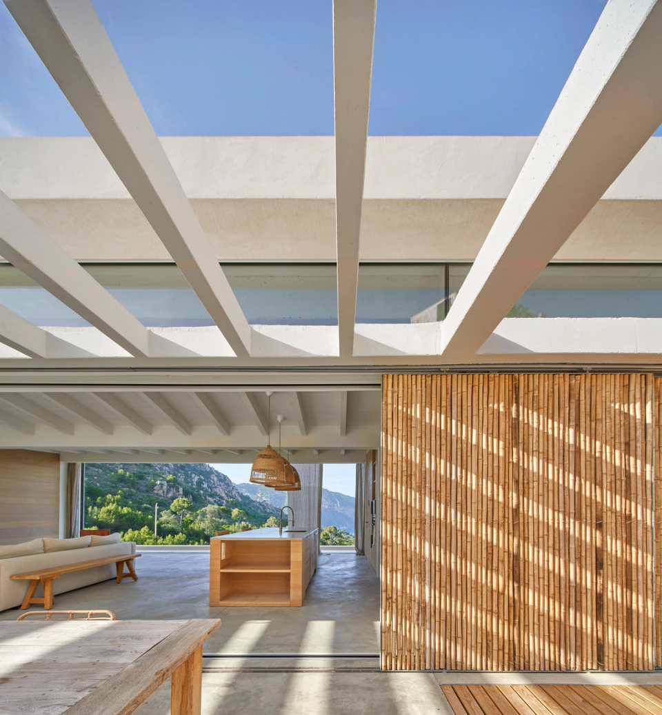 This single-family home stands out for its contemporary Mediterranean architecture, based on transparency, natural light, and a constant connection with the landscape. The structural pergola creates an elegant play of shadows over the interior and exterior spaces, providing depth and climatic comfort throughout the day. Natural wood enclosures and large sliding openings transform the boundaries of the house, integrating the living room, kitchen, and terrace into one fluid and open space. This single-family home combines noble materials, minimalist lines, and a direct relationship with the natural surroundings to create serene, functional architecture fully connected to outdoor living.