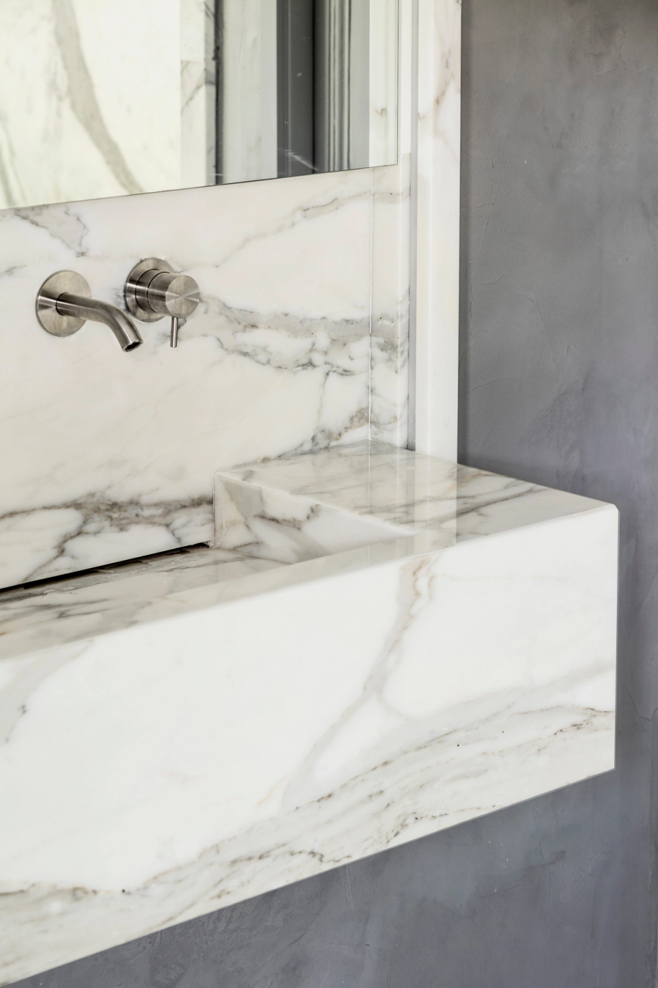 This interior renovation reflects a refined minimalist aesthetic through the use of white marble with natural veining, bringing visual continuity and a sense of exclusivity to the bathroom. The suspended straight-lined sink combined with built-in metallic fixtures creates a contemporary and elegant atmosphere, while the gray-toned walls add contrast and sophistication.