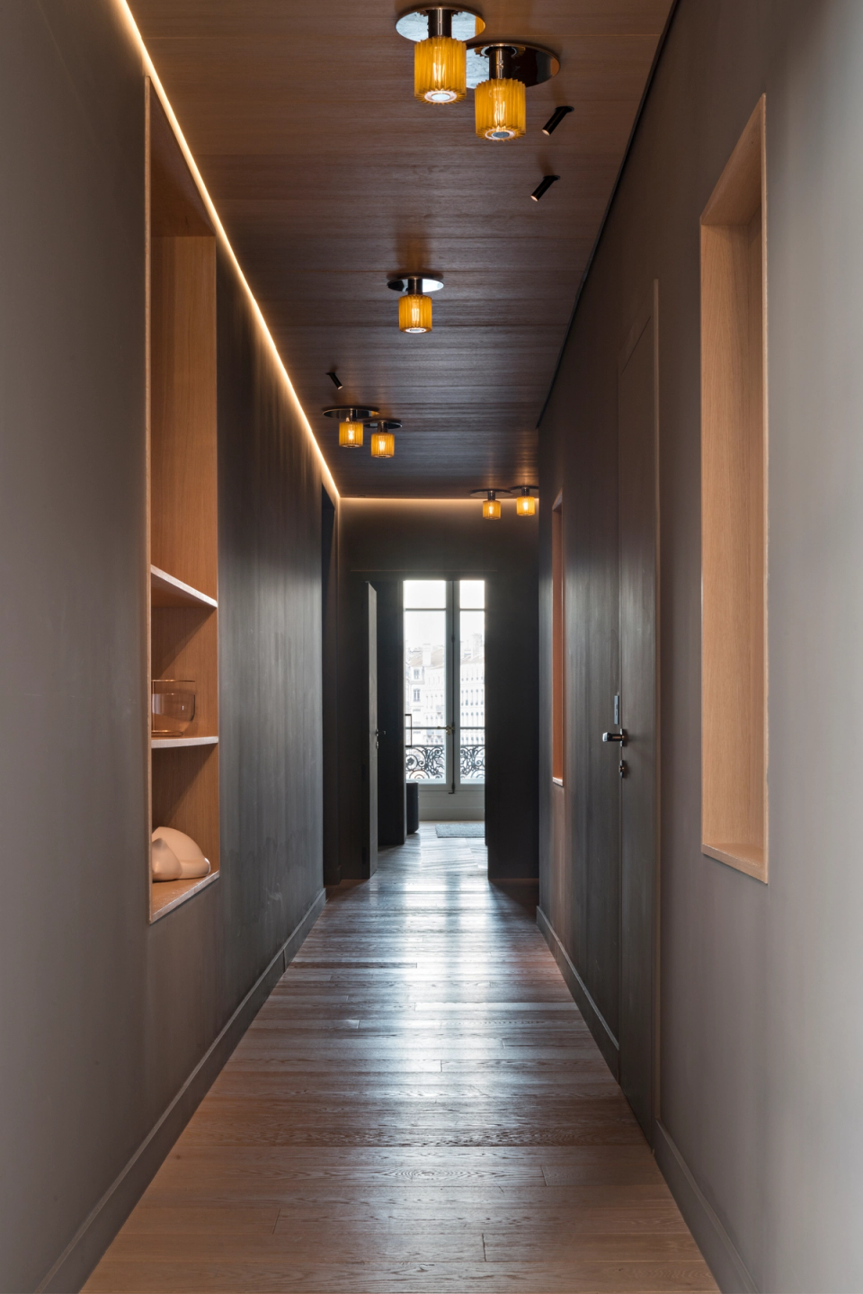 The interior renovation of this hallway transforms a circulation area into a space with its own identity, thanks to a carefully curated composition of light, materials, and proportions. Dark finishes and the continuous wooden ceiling add depth and elegance, while integrated linear lighting and decorative fixtures create a warm and sophisticated atmosphere. Illuminated niches and clean lines reinforce the contemporary language of the project, creating a visually coherent and immersive pathway.