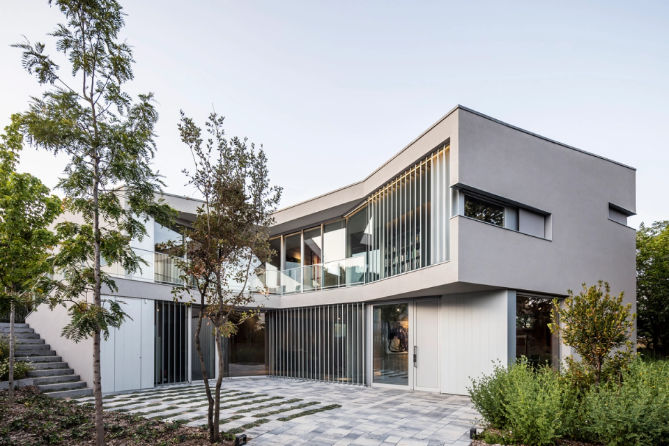 Detached single-family home with a contemporary architectural style featuring clean lines and geometric volumes that create an elegant and sophisticated appearance. The façade combines light finishes, large glazed surfaces, and vertical slats that provide rhythm, privacy, and control of natural light. The outdoor access area, surrounded by vegetation and permeable paving, reinforces the integration of the house with its surroundings and enhances the sense of spaciousness, brightness, and connection between interior and exterior spaces.