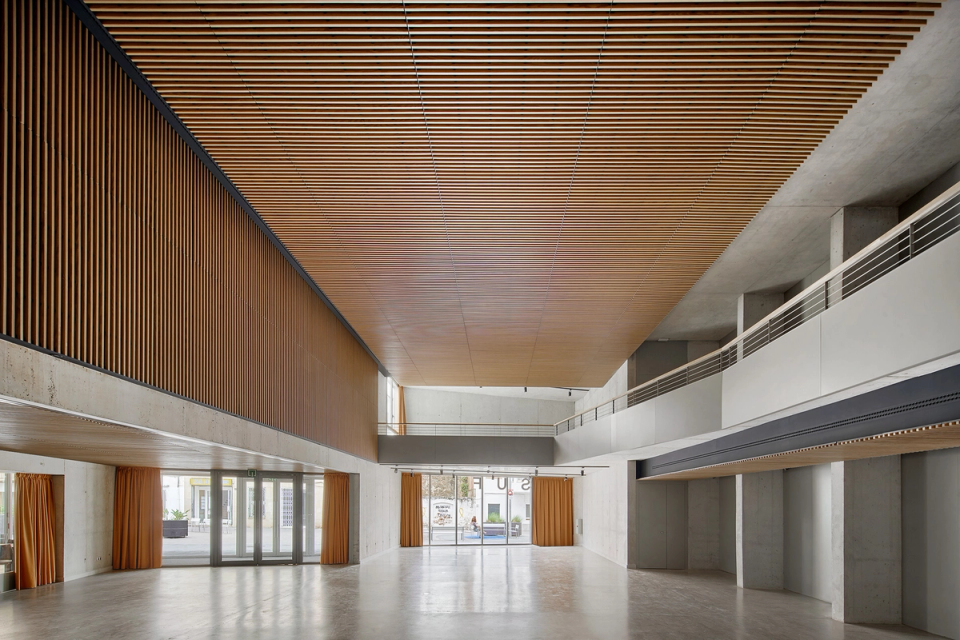 Interior detail of a cultural center with contemporary architecture distinguished by the use of materials and spatial quality. Wooden slats on ceilings and cladding provide acoustic and visual warmth, while exposed concrete reinforces the sober and functional character of the space. The open configuration and the height of the lobby allow the cultural center to adapt to exhibitions, stage activities, and community events, creating a modern, flexible environment designed for public use.