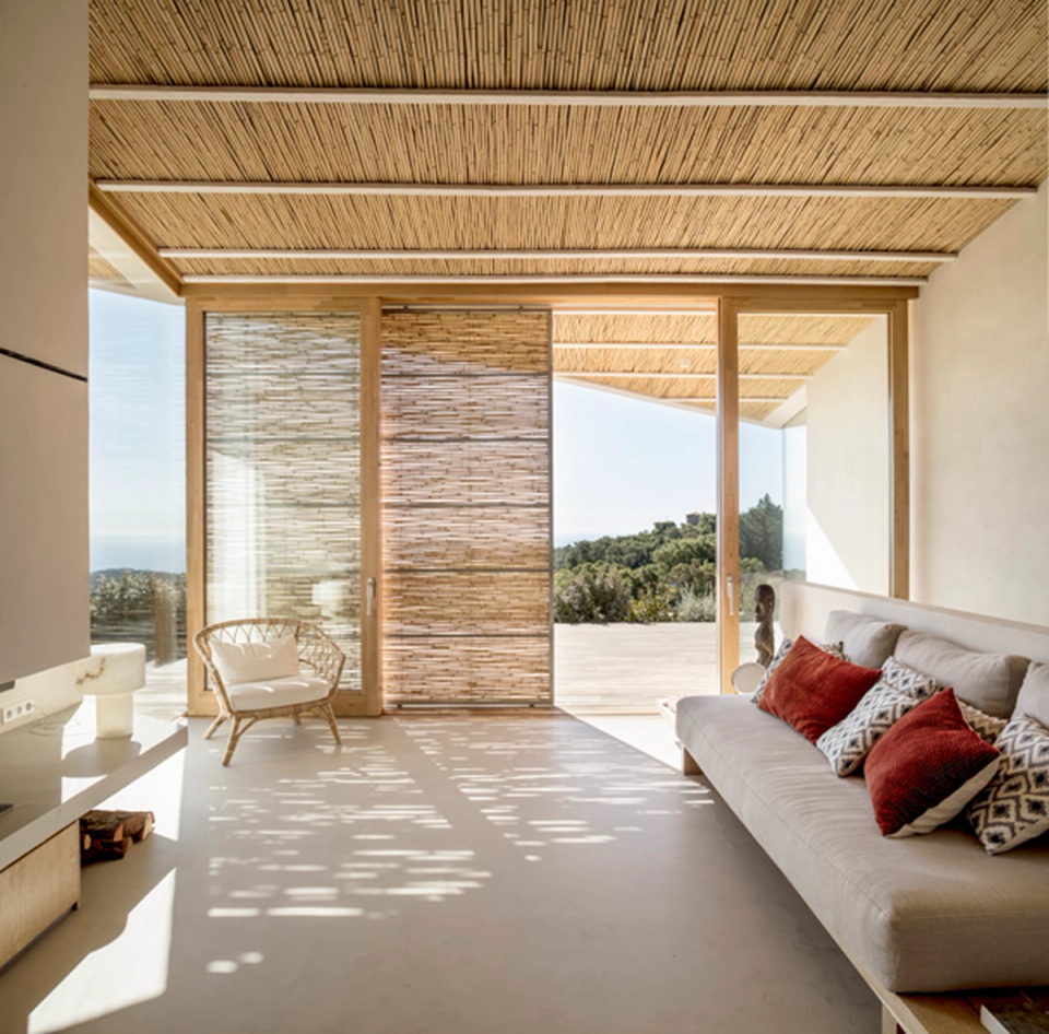 Living room of a detached house with warm minimalist interior design combining natural materials, integrated wooden furniture and a contemporary fireplace as the central feature of the space. Ceilings clad with natural fibers and vertical openings provide texture, natural light and spaciousness, creating a welcoming atmosphere connected with the Mediterranean architecture of the home.