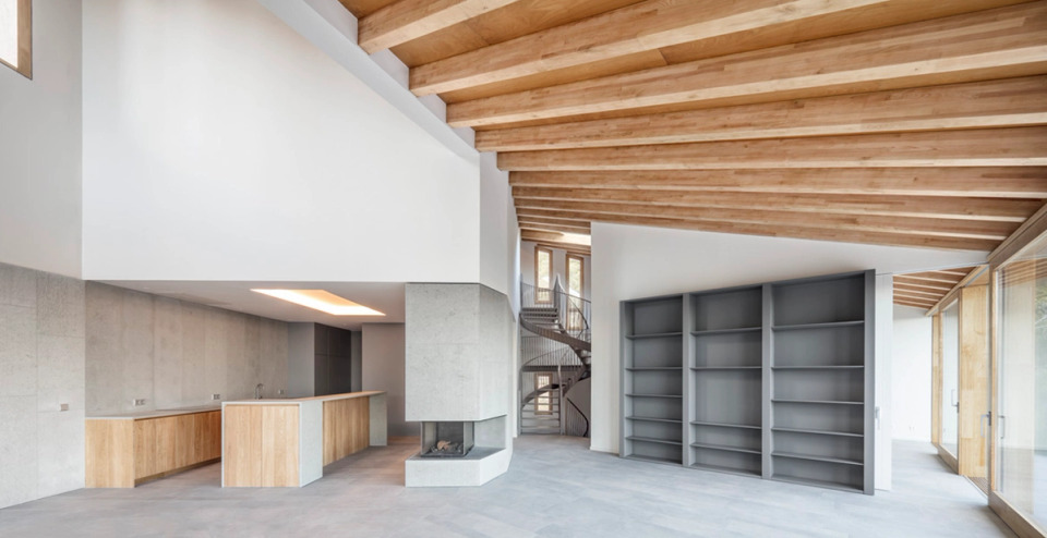 Open-plan living area of a detached single-family home with a fluid and contemporary layout integrating kitchen, living room, and circulation spaces into one spacious and bright environment. Ceilings with exposed wooden beams, neutral finishes, and large exterior openings provide warmth and connection with the surroundings, while the central fireplace and sculptural staircase in the background reinforce the home’s unique architectural character.