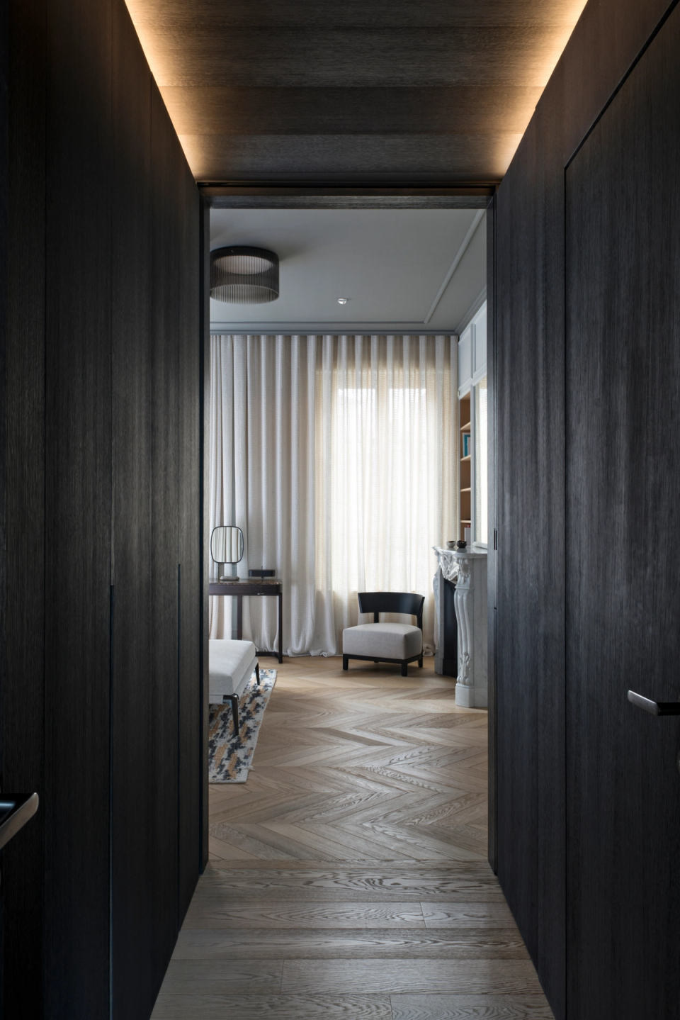 The interior renovation of this home highlights the connection between spaces through a carefully curated combination of textures, light, and noble materials. The hallway clad in dark wood creates an intimate and sophisticated transition toward the main rooms, where herringbone parquet flooring, classical moldings, and soft textiles bring warmth and elegance. Integrated indirect lighting reinforces the depth of the spaces and creates a serene, contemporary, and timeless atmosphere.