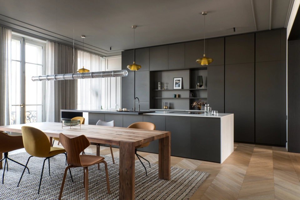 The interior renovation of this open kitchen redefines the social space of the home with an elegant, functional, and contemporary proposal. Dark-toned furniture adds depth and sophistication, while the natural wood flooring and dining table balance the composition with warmth and texture. The large central island and the clean lines of the furniture enhance the feeling of order and spaciousness, complemented by decorative lighting that reinforces the exclusive and refined character of the space.