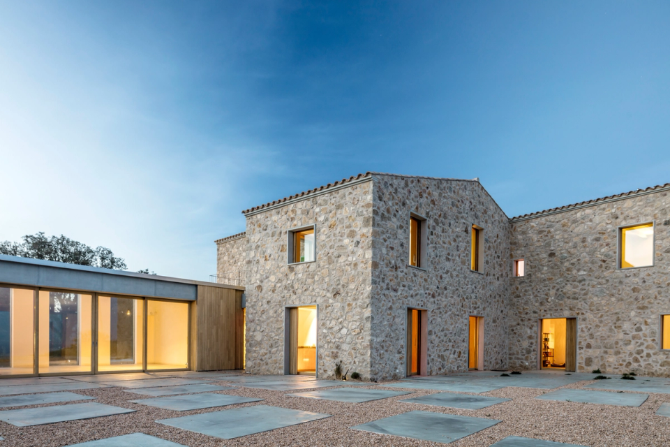Exterior view of a comprehensive renovation that recovers the essence of traditional architecture while integrating contemporary volumes and natural materials. The home adapts sensitively to the rural landscape, combining restored stone façades, wooden carpentry, and large openings that connect the interior spaces with the surroundings. Warm lighting and respectful placement on the terrain reinforce the serene and timeless character of this full renovation, conceived to merge heritage, comfort, and contemporary architecture.