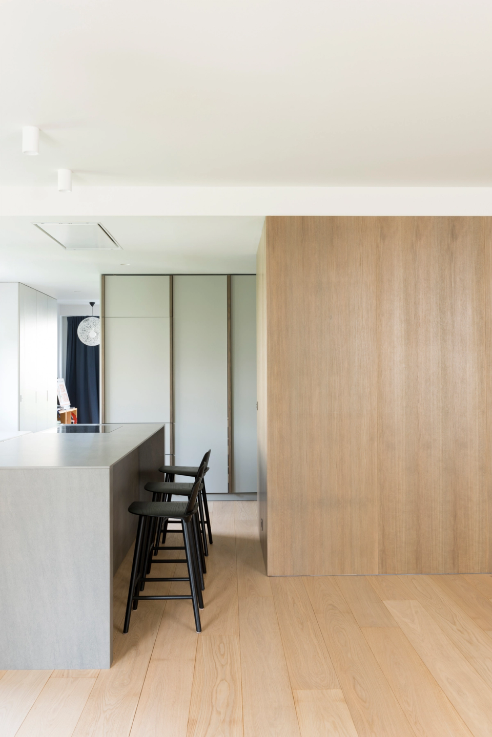 Interior renovation of a contemporary home with open spaces and a warm minimalist aesthetic. The kitchen, integrated into the living-dining area, stands out for its large stone-gray central island, custom furniture, and natural wood finishes that provide continuity and visual comfort. Natural light generously enters through large windows, enhancing the feeling of spaciousness and serenity. The project combines clean lines, noble materials, and contemporary furniture to create an elegant, functional, and timeless atmosphere.