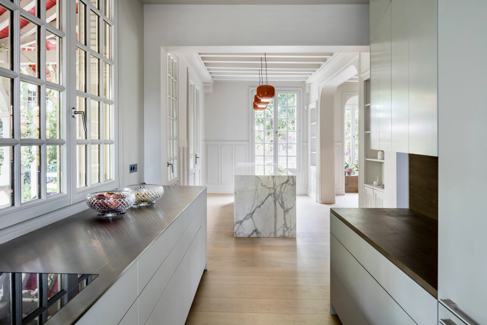 Interior renovation of a contemporary kitchen integrated into a classical-style residence, where natural light and visual continuity between spaces take center stage. Minimalist cabinetry lines, neutral-toned finishes, and a central marble island contrast with original moldings and large openings, creating an elegant balance between tradition and modernity.