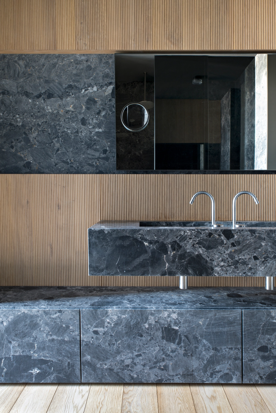 The interior renovation of this bathroom stands out for the combination of dark natural stone and fluted wood cladding, creating an elegant, contemporary composition full of personality. The monolithic furniture design enhances the sense of continuity and sophistication, while the minimalist lines of the fixtures and mirrors reinforce a clean and balanced aesthetic. Contrasts between mineral textures and warm materials add visual depth and transform the space into an exclusive environment designed to convey comfort, functionality, and architectural refinement.