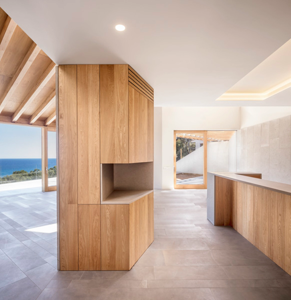 Open kitchen in a detached single-family home with natural wood cabinetry and a minimalist layout integrated into a spacious and bright environment. Large openings visually connect the interior with the terrace and sea views, while the combination of warm materials, clean lines, and natural light reinforces the contemporary and welcoming character of the home.