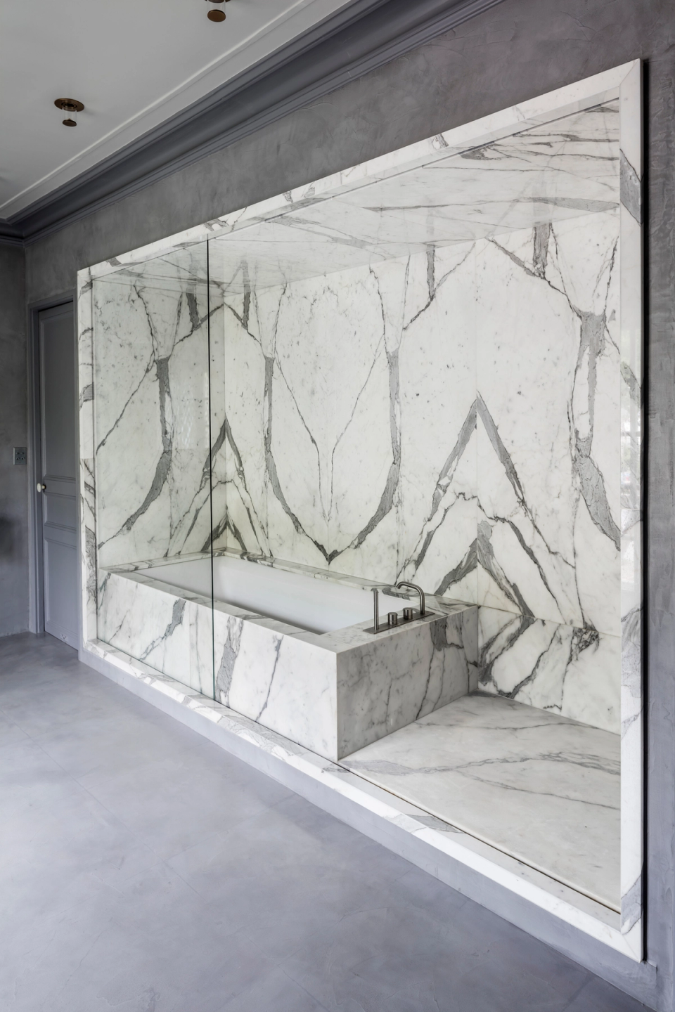 This interior renovation embraces an elegant and sophisticated design where large-format marble becomes the absolute protagonist of the bathroom, creating a sense of continuity and contemporary luxury. The gray tones of the walls and doors provide depth and visual balance, while integrated lighting highlights the natural veining of the stone and enhances the spaciousness of the room with an exclusive and minimalist result.