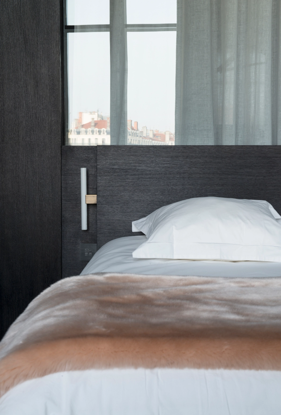 The interior renovation of this bedroom stands out for its minimalist and sophisticated aesthetic, where noble materials and soft textures create an intimate and elegant atmosphere. The dark wood-clad headboard subtly integrates lighting, adding visual depth and reinforcing the contemporary character of the space. Natural light filtered through lightweight curtains enhances the sense of calm and comfort, while the combination of neutral tones and warm finishes transforms the room into a welcoming and refined resting area.