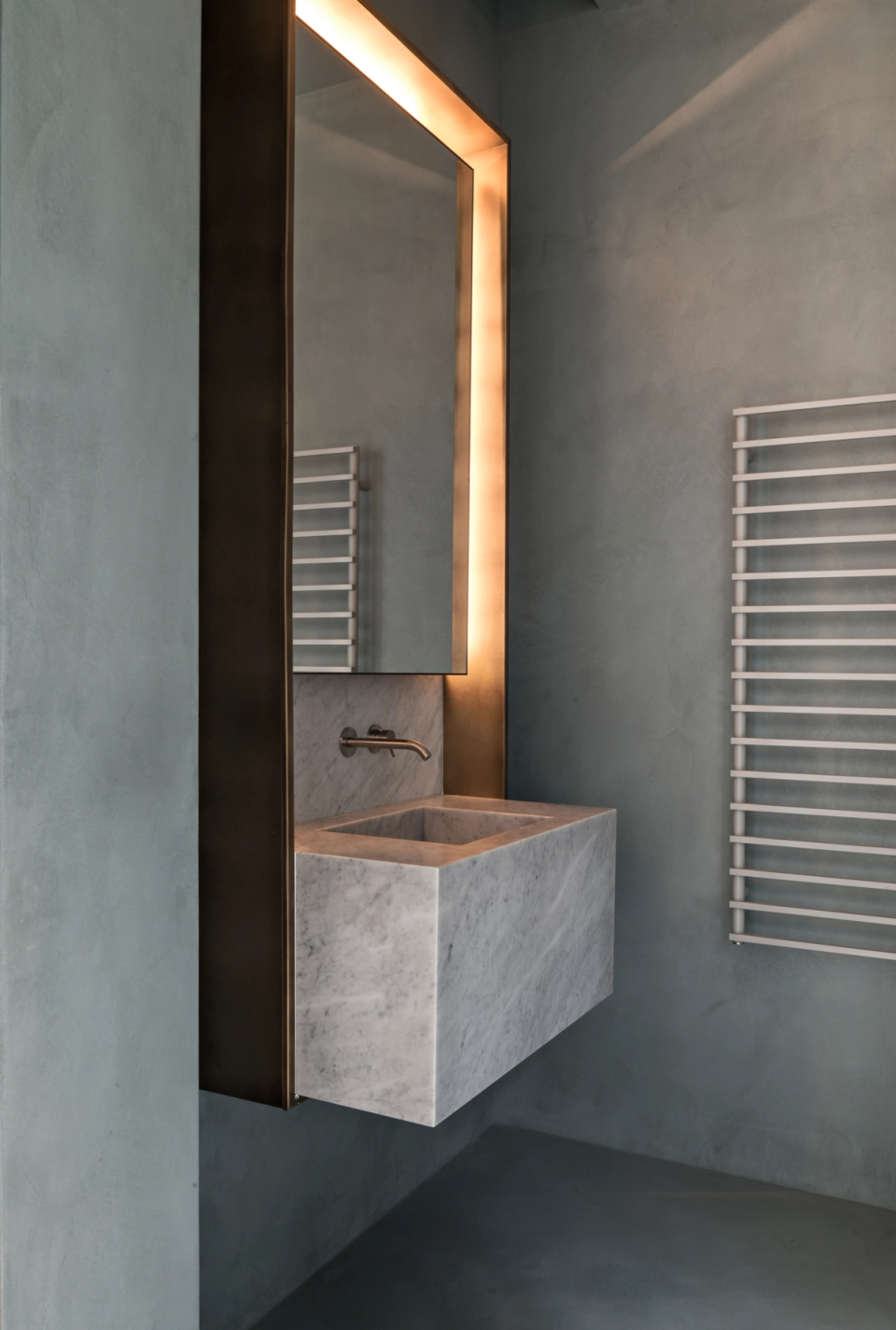 The interior renovation of this bathroom embraces a refined and timeless aesthetic, where natural stone becomes the absolute protagonist. The suspended monolithic sink, combined with continuous gray-toned surfaces and integrated mirror lighting, creates a serene and sophisticated atmosphere. Minimalist finishes and the subtle combination of textures add visual depth and reinforce the elegant and contemporary character of the space.