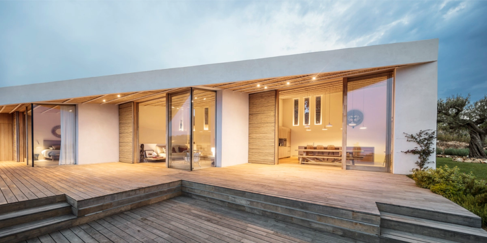 Exterior view of a detached house at sunset, featuring contemporary architecture with clean lines and large glass openings that reveal the warmth of the interior spaces. The wooden terrace, integrated lighting beneath the pitched roof and the seamless connection between inside and outside create an elegant and welcoming atmosphere perfectly integrated into the surrounding natural landscape.