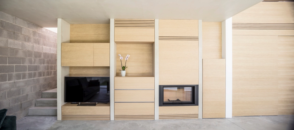 Integrated natural wood furniture organizes the space through a clean and functional composition, incorporating storage, fireplace, and household equipment into a single continuous element. The combination of warm textures and neutral materials reinforces the restrained and welcoming character of the interior.