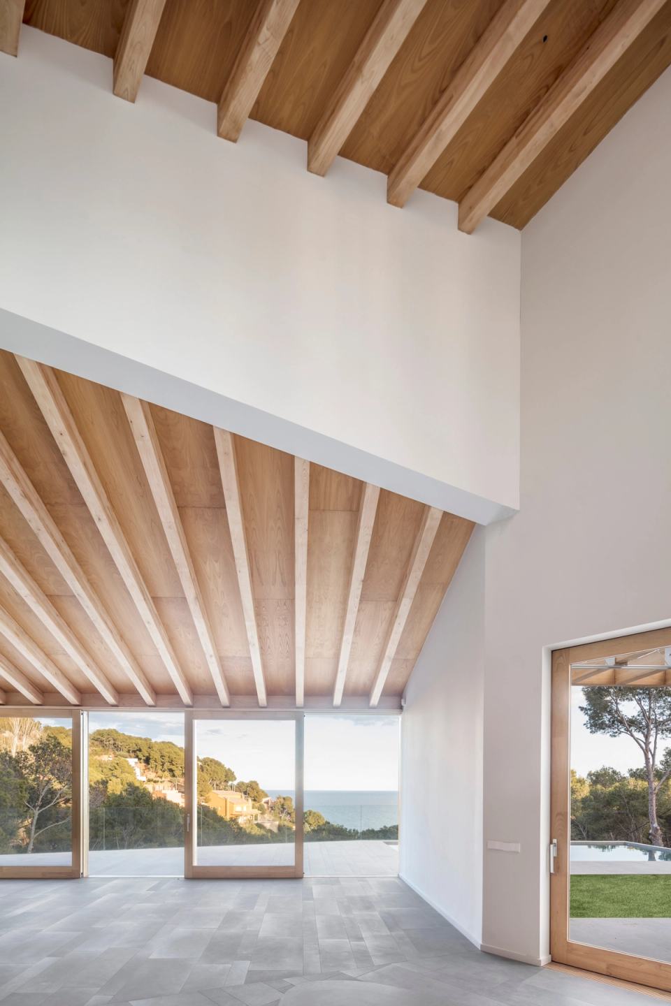Living room of a detached single-family home with large glass enclosures that fully open the house to the surrounding landscape and sea views. Ceilings with exposed wooden structures provide warmth and rhythm to a spacious, bright space connected to nature, enhancing the fluid relationship between interior, terrace, and Mediterranean surroundings.