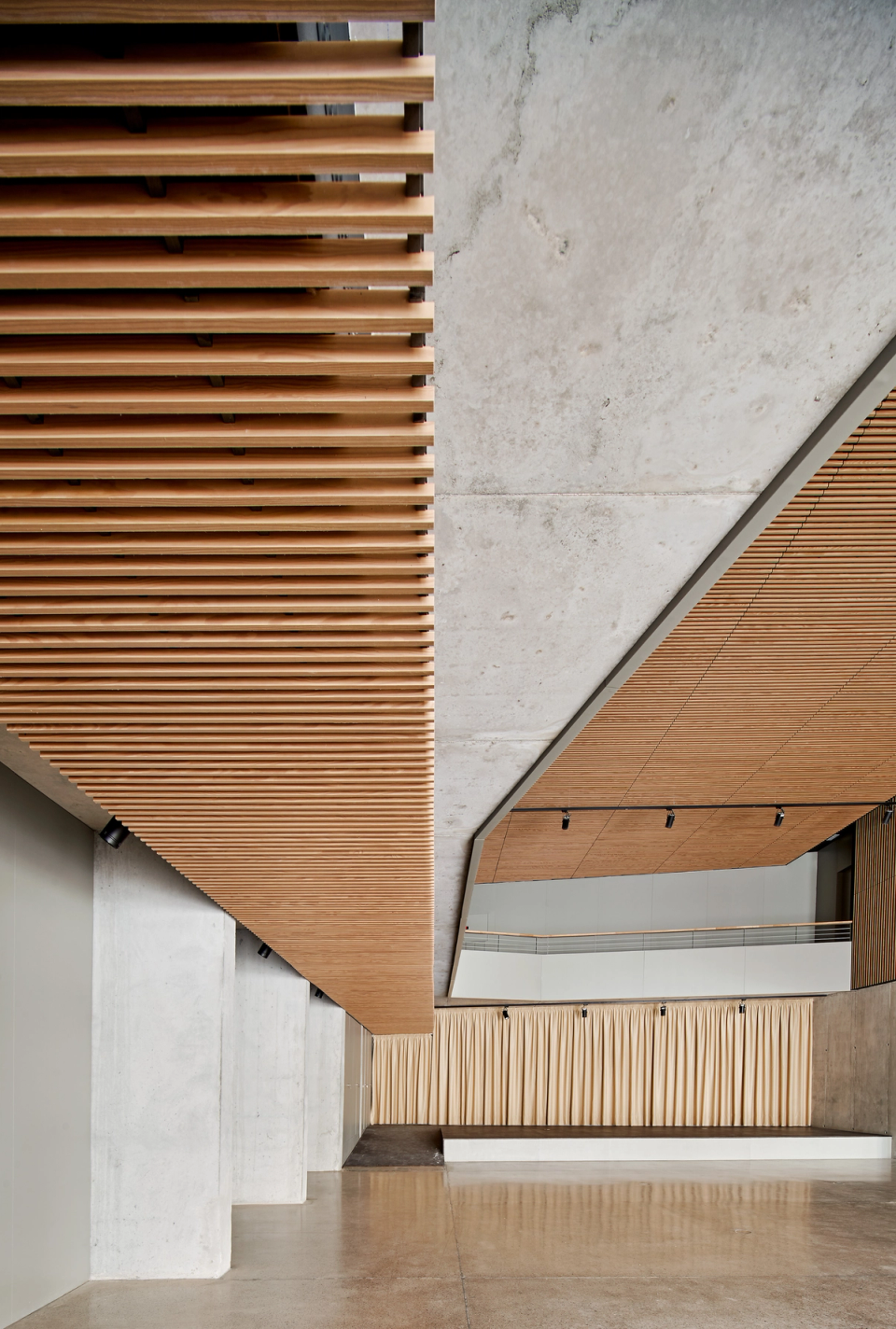 Central space of a cultural center with an interior architecture defined by functionality, light, and material quality. Large wooden slat ceilings provide acoustic comfort and a contemporary image, while upper walkways and the integrated stage allow the space to adapt to multiple cultural and social activities. The combination of exposed concrete, natural wood, and generous dimensions creates a spacious, flexible environment designed to turn the cultural center into a community meeting point.