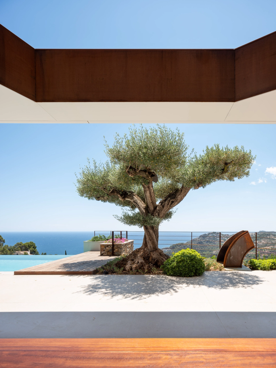 Outdoor space of a luxury detached single-family home where architecture, landscape, and nature merge into an elegant and balanced composition. The century-old olive tree becomes the visual centerpiece of the space, framed by contemporary architectural lines, noble materials, and open views of the Mediterranean Sea. The integration of corten steel, wood, and minimalist surfaces reinforces the exclusive character of the home, creating a serene and sophisticated atmosphere that enhances the connection between interior, exterior, and the natural surroundings.