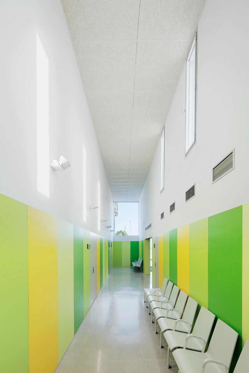 Interior corridor of the Sant Vicenç Health Center conceived as a bright, functional, and easily recognizable space. Vertical bands in green and yellow tones provide identity and help orient circulation within the center, while natural light entering through upper openings creates a welcoming and comfortable atmosphere for users.