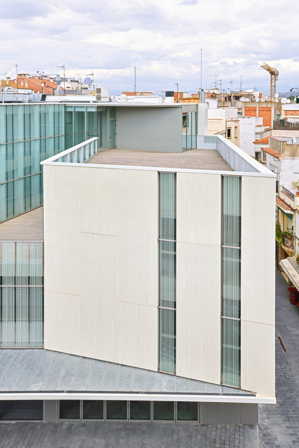 Exterior detail of a contemporary cultural center with a façade clad in light finishes and vertical openings that emphasize its elegant and minimalist volumetry. The combination of opaque surfaces and large glass enclosures creates a bright and balanced architecture integrated into the urban environment. The terraces and open spaces encourage the relationship between interior and exterior, providing functionality and spatial quality to this cultural facility.