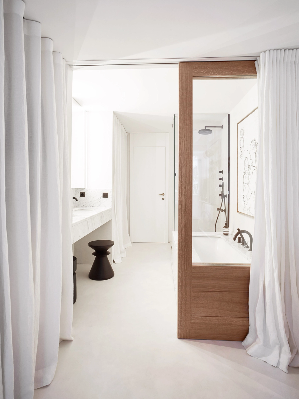 The full renovation of the bathroom and suite embraces an open and sophisticated layout where the different spaces connect fluidly through textile curtains and natural wood elements. The combination of white surfaces, light stone and matte black details creates an elegant and contemporary contrast that reinforces the sense of serenity and exclusivity. The bathtub, integrated within a wooden volume, becomes a central feature of the project, bringing warmth and personality to the space. Natural light, noble materials and minimalist design transform this area into an intimate and relaxing environment designed to enhance everyday comfort and wellbeing.