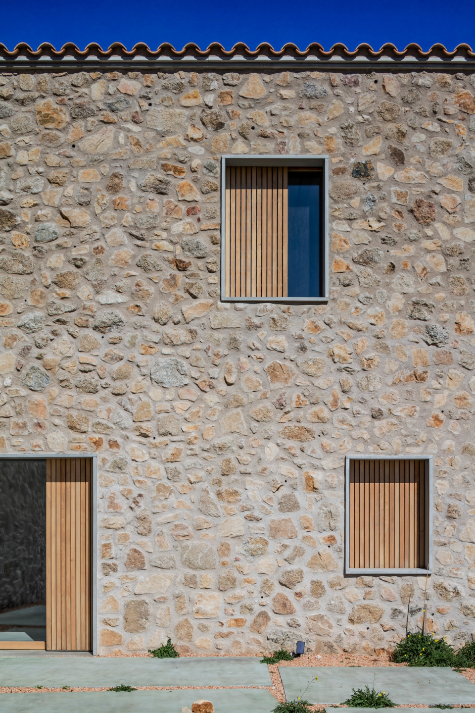 Façade detail of a comprehensive farmhouse renovation project featuring restored natural stone walls and new wooden carpentry elements that introduce a contemporary and elegant language. The architectural intervention respects the original materials of the traditional construction, highlighting the texture and authenticity of the stone while incorporating modern solutions that improve efficiency, natural light, and the functionality of the home. This full renovation stands out for the attention to finishes, the harmony between tradition and modernity, and a sober architecture integrated into the rural environment.