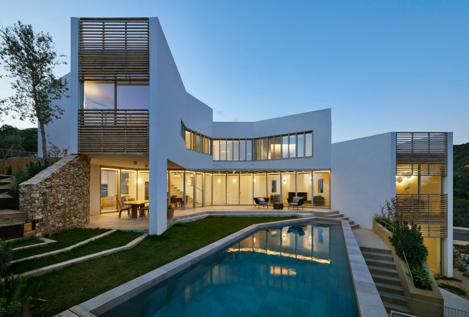Exterior of a contemporary detached single-family home with swimming pool, large glazed surfaces, and geometric volumes integrated into the terrain. The project combines modern design, natural light, and open spaces to create a fluid connection between interior and exterior.