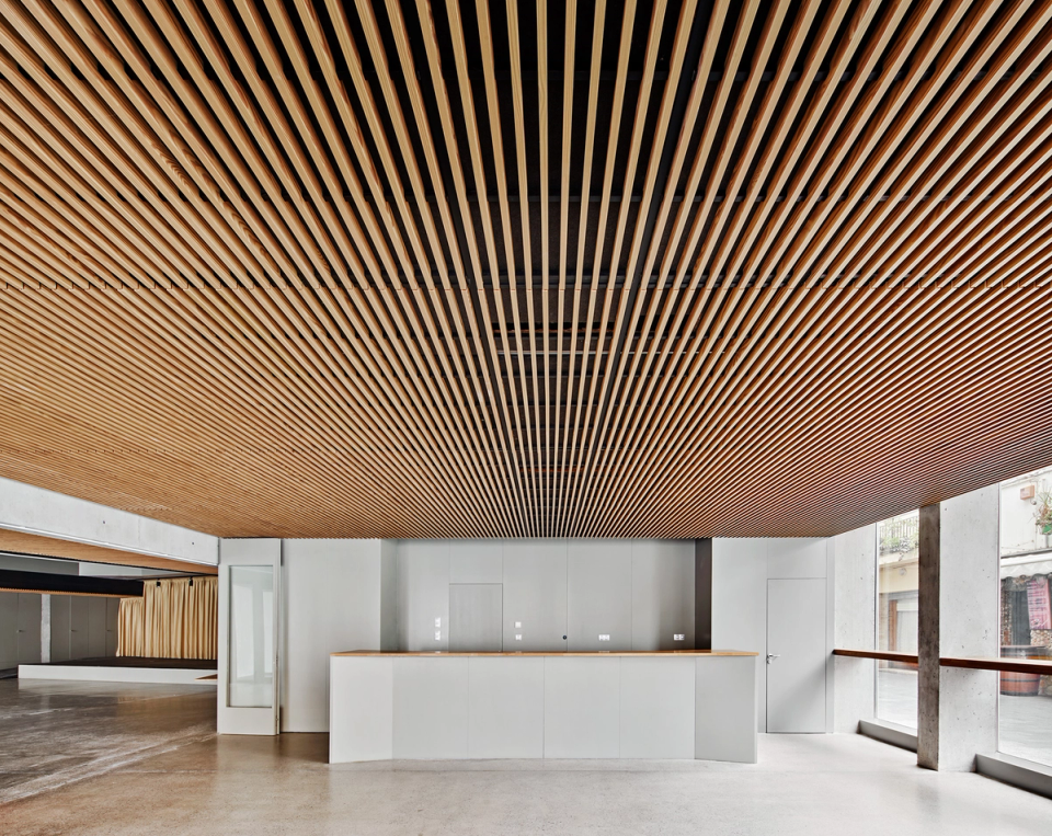 Interior walkway of a contemporary cultural center that visually connects the different levels of the building through a large open central space. Natural wood finishes on the ceiling and vertical cladding provide warmth and acoustic quality, while exposed concrete reinforces the modern and functional character of the architecture. This area of the cultural center has been conceived to encourage circulation, interaction between users, and flexibility of use in cultural and social activities.
