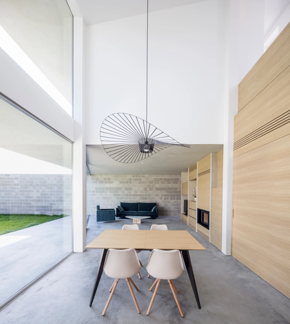 The interior space stands out for its spaciousness and brightness, enhanced by double-height ceilings and large glass enclosures. The combination of polished concrete, natural wood, and neutral surfaces creates a warm and minimalist atmosphere in continuous visual connection with the outdoor garden.
