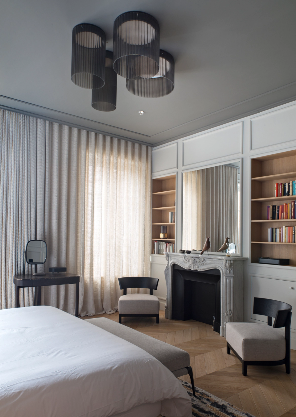 The interior renovation of this bedroom combines classical elegance and contemporary design to create a warm, balanced, and sophisticated space. Original moldings, the ornamental fireplace, and herringbone flooring interact with refined-lined furniture and sculptural decorative lighting. Linen curtains softly filter natural light, while wood finishes and neutral tones bring comfort and serenity to an environment designed for rest and well-being.